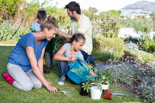 A family working together in a well-maintained garden, featuring a lush green lawn in the foreground and vibrant flower beds bordering the grassy area. The scene shows a woman kneeling on the lawn, planting or tending to the grass and garden plants using a small gardening fork, while a young girl sits nearby with a deep blue backpack, possibly involved in planting or watering. Behind them, a man is crouched down, supervising or assisting with gardening tasks, with two children standing close by, one wearing a striped shirt. The garden includes a variety of flowering plants, shrubs, and small trees, with some potted plants and garden tools such as a watering can and trowel resting on the grass. The outdoor space appears sunny with natural daylight, surrounded by a hedge and flowering bushes, demonstrating a tidy and inviting garden environment typical of residential landscaping in Newham, which Lawn Mowing Newham services. The garden layout emphasizes a spacious, well-kept lawn bordered by colorful plantings, suitable for outdoor family activities and gardening maintenance. This scene highlights routine outdoor garden care, reflecting professional lawn and garden services in the local area.
