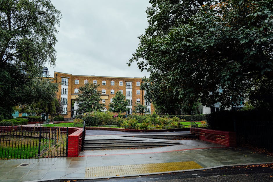 A landscaped communal garden in East Ham, featuring a paved entrance with a tactile yellow safety strip and a wheelchair ramp. The garden is bordered by black metal fencing with red brick posts, leadi