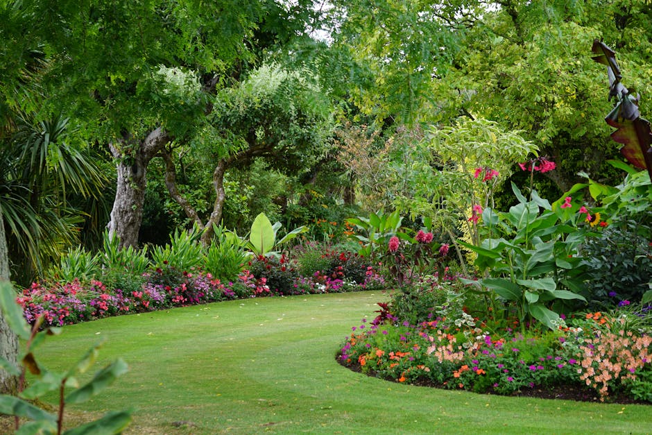 A lush, well-maintained garden scene featuring a neatly clipped green lawn with a gentle curve, bordered by vibrant flower beds filled with colorful blooms including pinks, oranges, and purples. Tall 