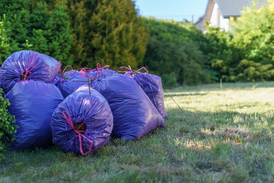 A row of seven purple gardening waste bags secured with pink ties, situated on a well-maintained lawn in a garden. The bags are filled with yard debris and are placed near a lush green hedge with dens